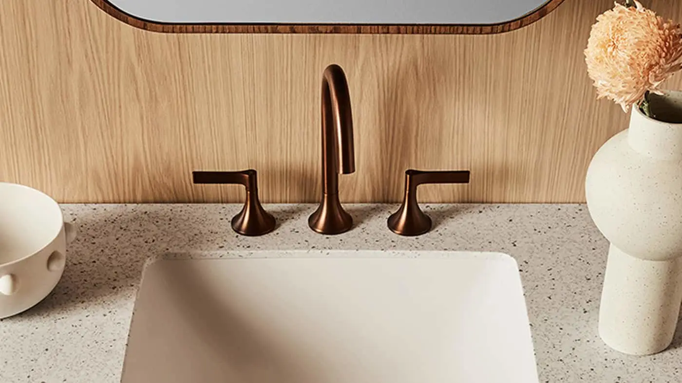 Modern bathroom sink with copper faucet, speckled countertop, wooden backsplash, and small white vase with dried flower.