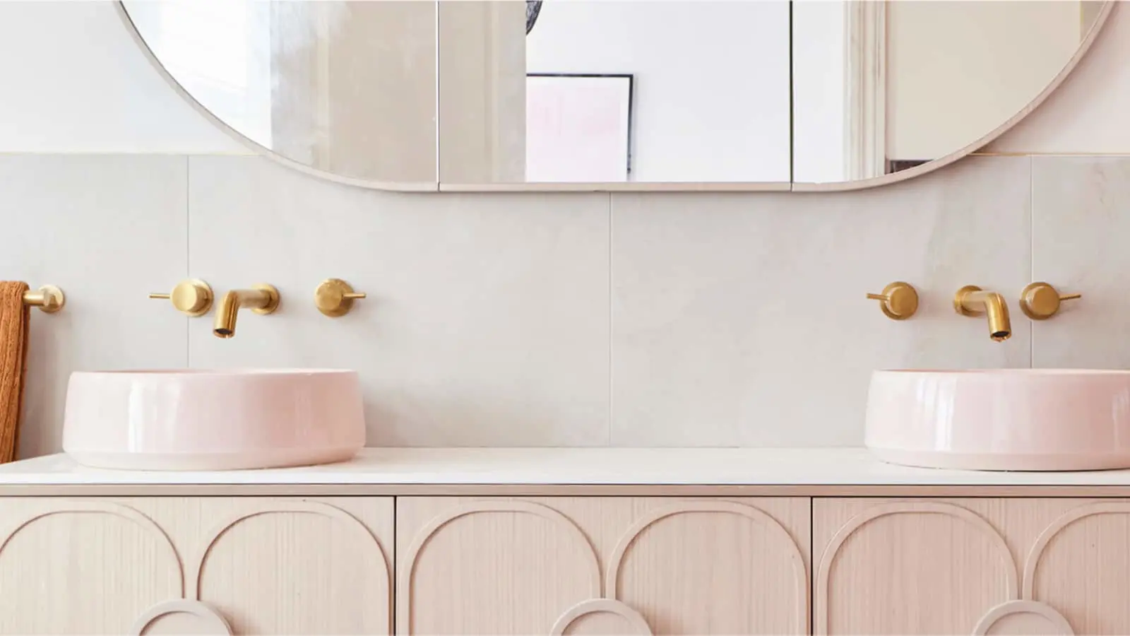 Modern bathroom with dual pink vessel sinks, brass wall-mounted faucets, and a wide mirror above light wood cabinetry.