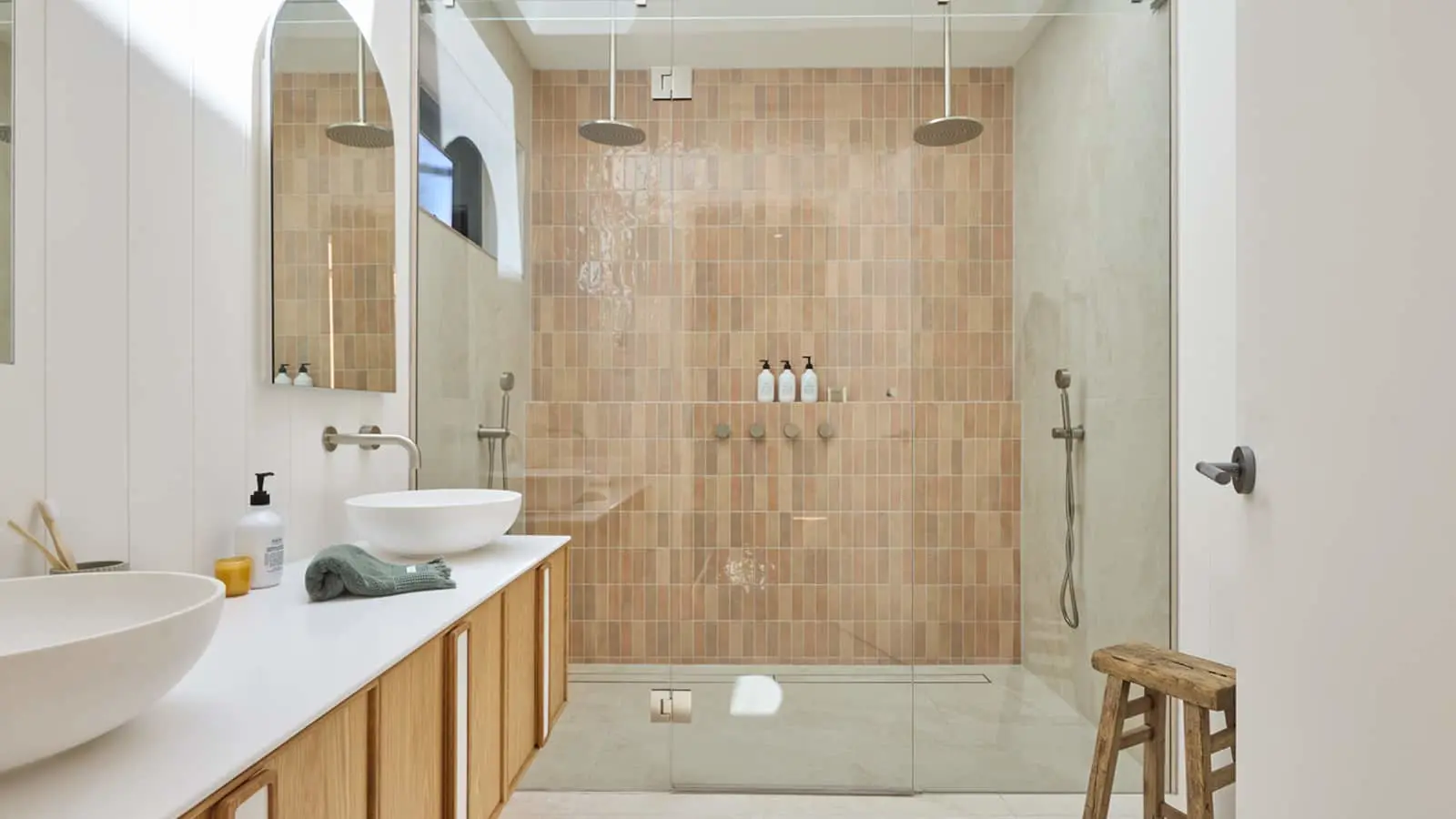 Modern bathroom with peach tile shower wall, white vessel sinks on wooden vanity, and glass shower enclosure.