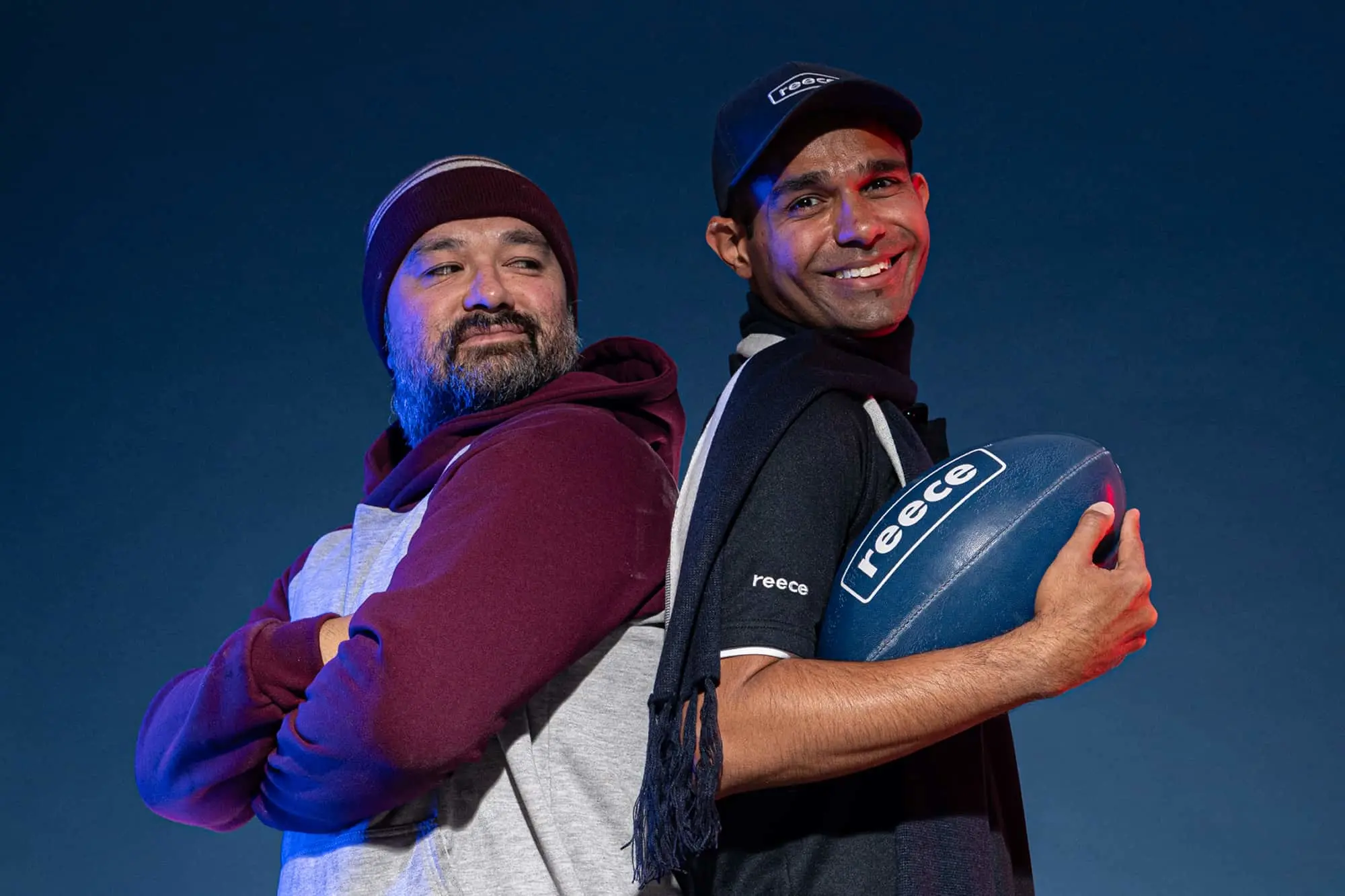 Two men standing back-to-back against blue background, one in maroon hoodie, other holding rugby ball wearing cap.