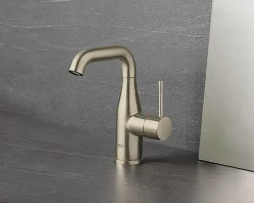 Modern brushed nickel bathroom faucet with curved spout and cylindrical handle against gray wall.
