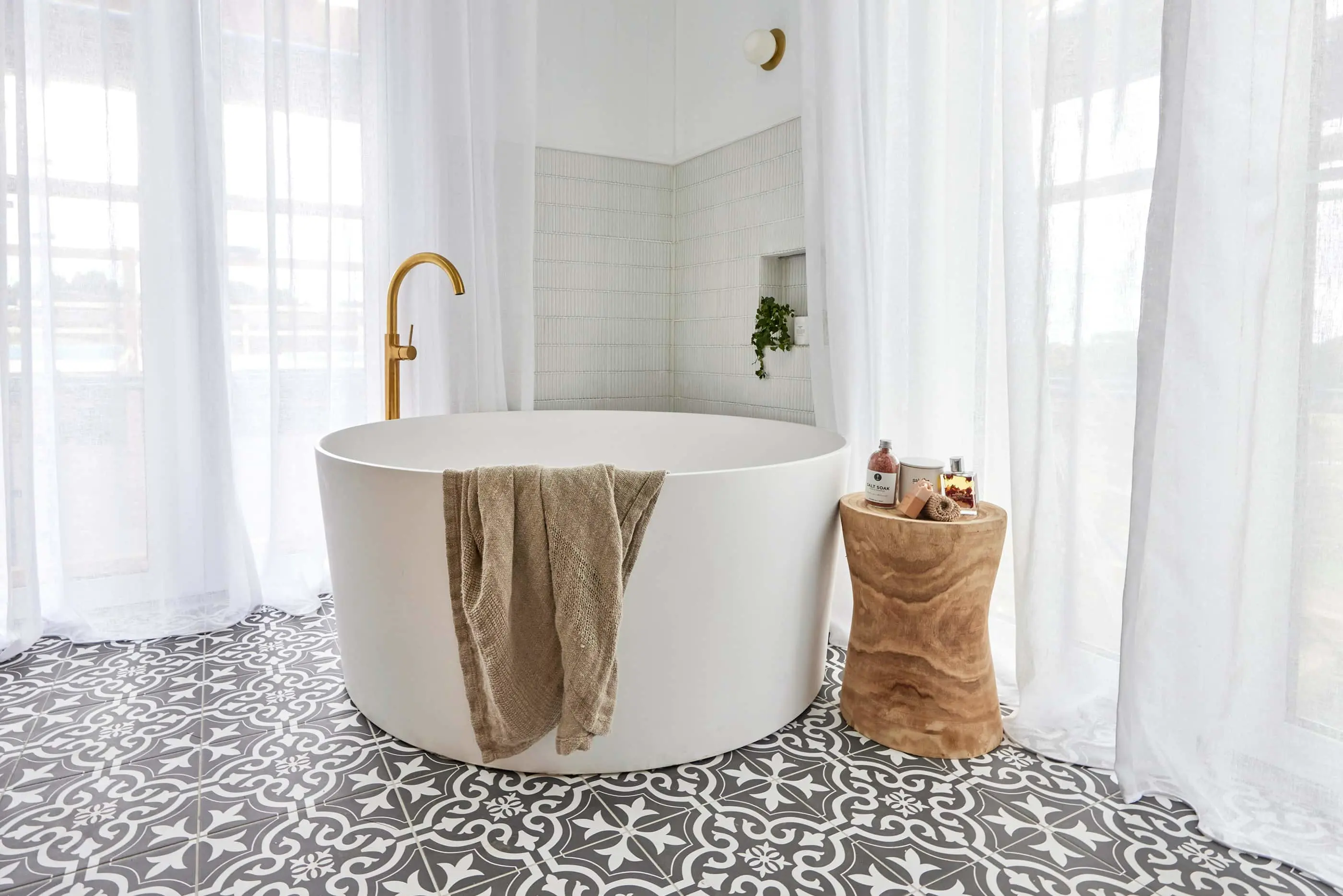 Modern white round bathtub with gold faucet, beige towel, wooden side table, patterned floor tiles, and sheer curtains.