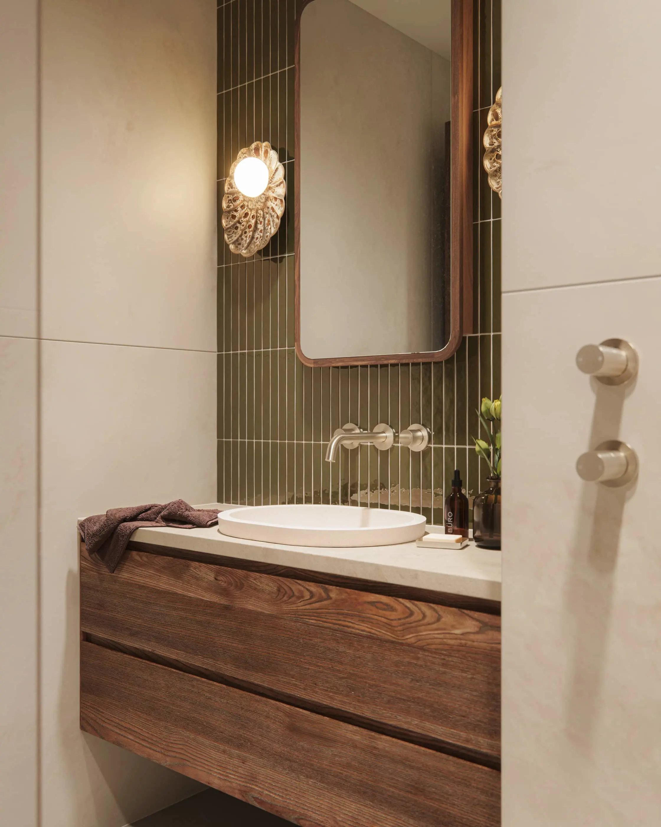 Modern bathroom with wooden vanity, green vertical tile, round sink, wall-mounted faucet, and shell-shaped sconce light.