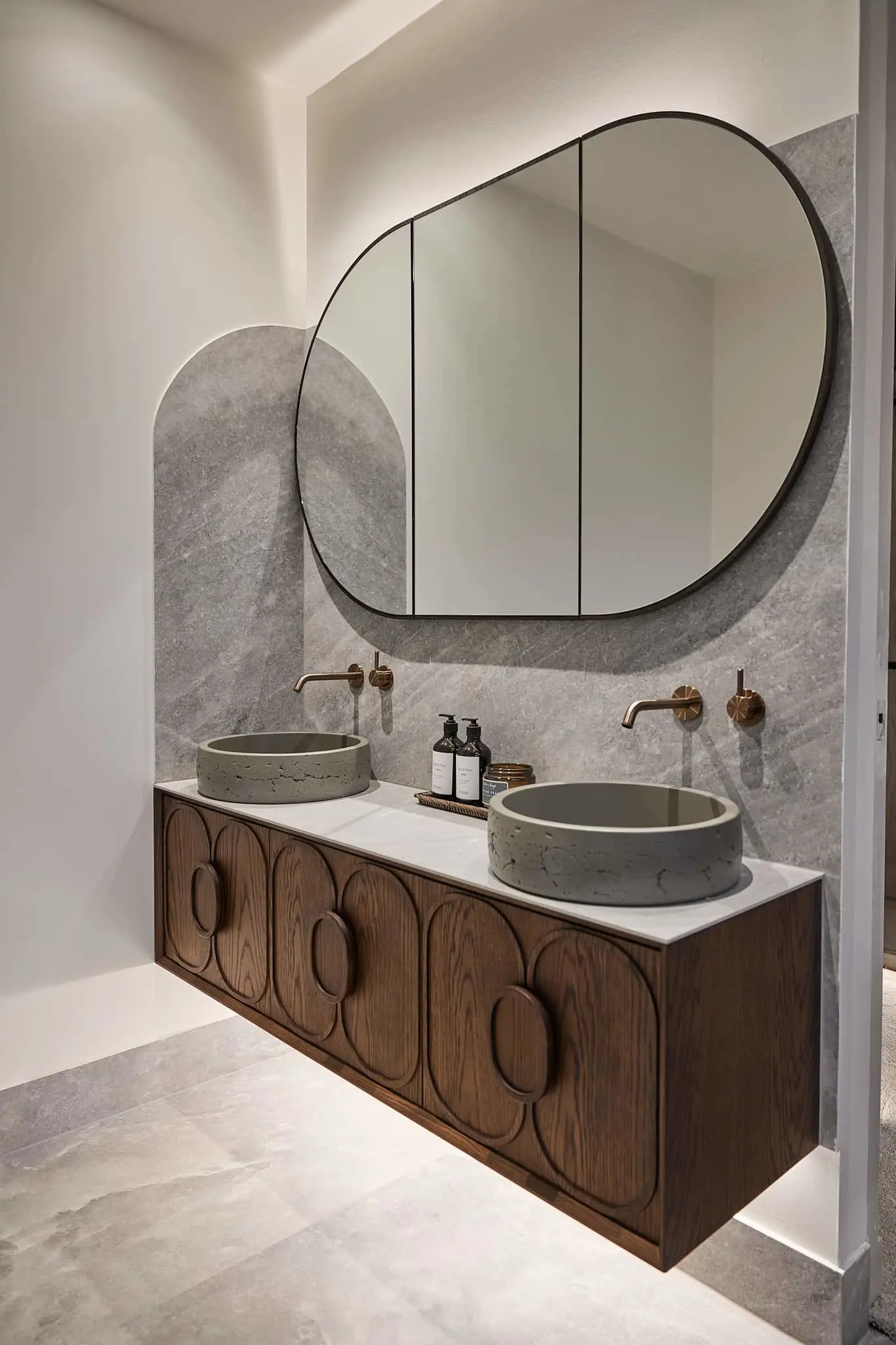 Modern bathroom with wooden vanity, two concrete vessel sinks, brass fixtures, and a large curved mirror above gray stone backsplash.