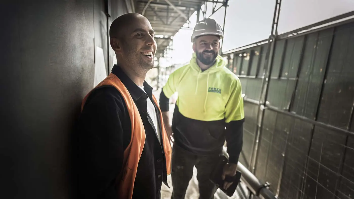 Two men smiling at a construction site, one wearing an orange vest, the other a yellow hoodie and white hard hat.