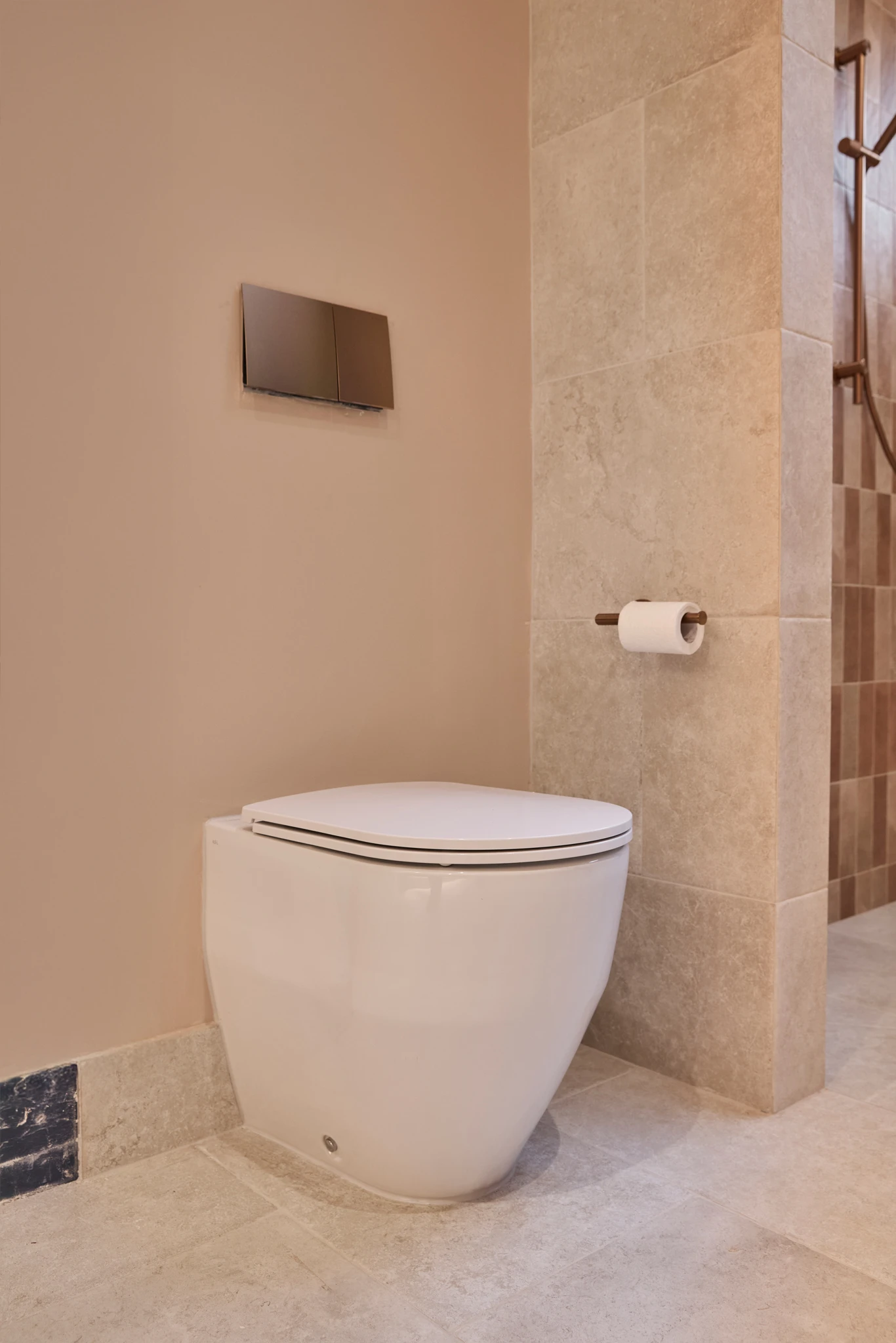 Modern bathroom with white toilet, beige tiled walls, wall-mounted flush panel, and toilet paper holder.