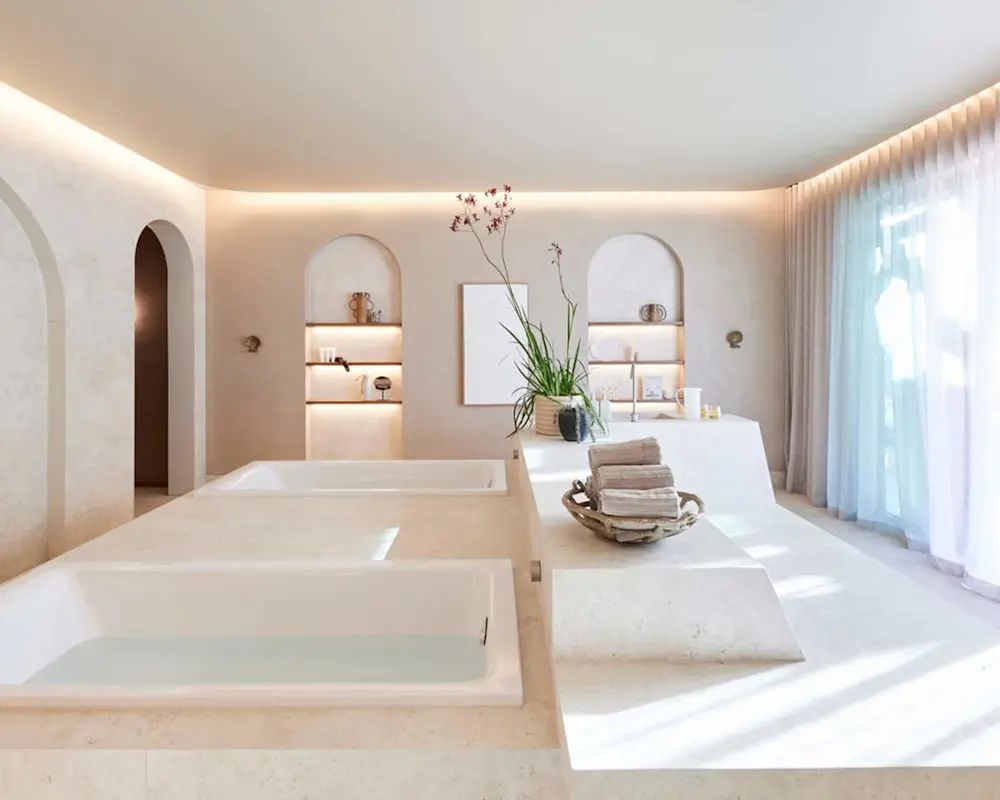 Minimalist spa bathroom with sunken tubs, arched alcoves, soft lighting, and cream-colored stone surfaces.