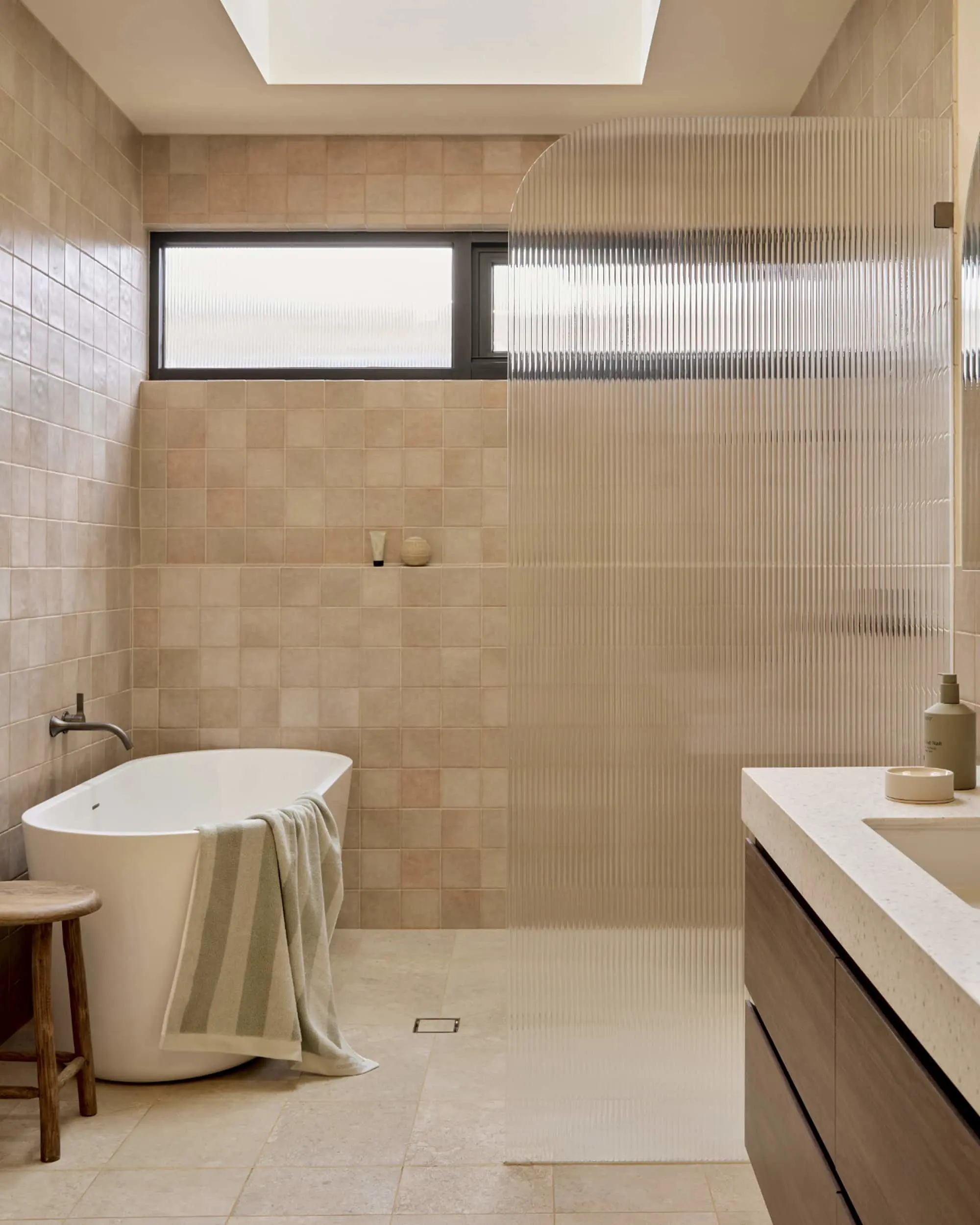 Modern bathroom with beige tiles, freestanding white tub, ribbed glass shower partition, and wooden vanity.
