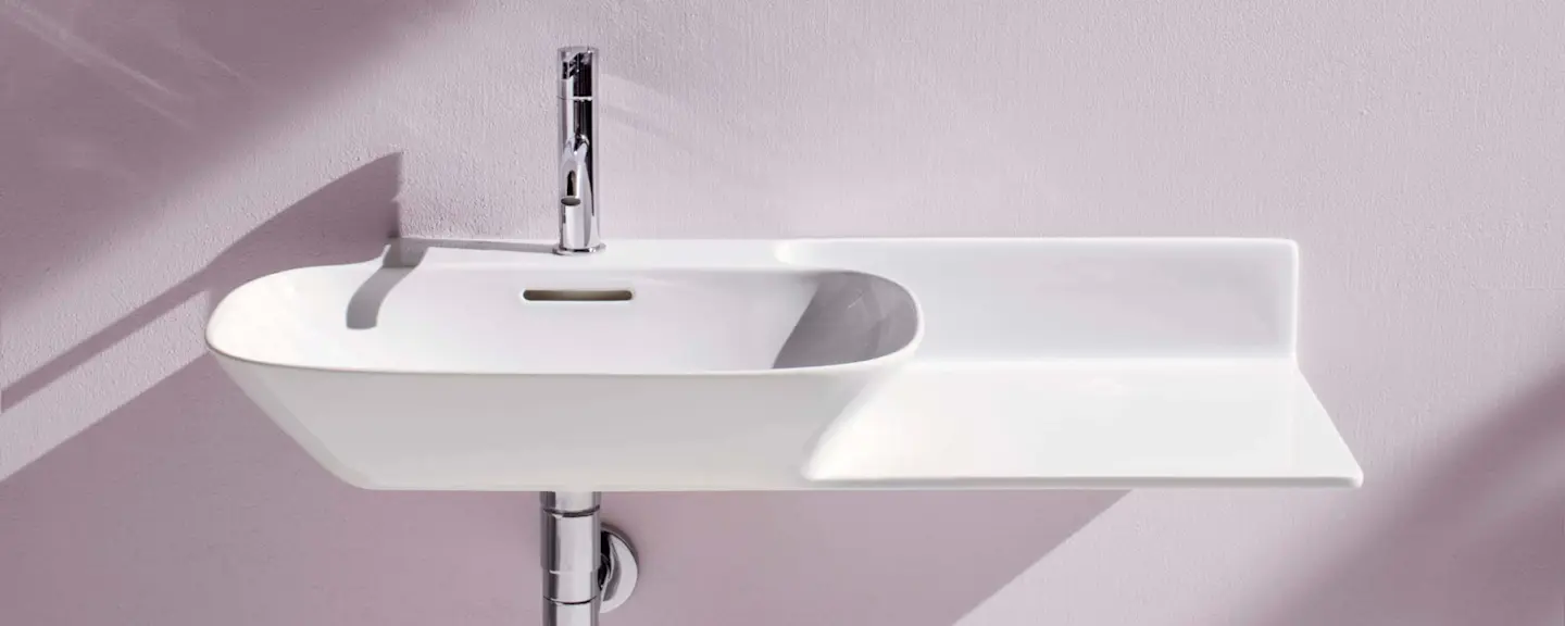 White wall-mounted bathroom sink with a chrome faucet and an extended flat surface on the right side, against a light pink wall.