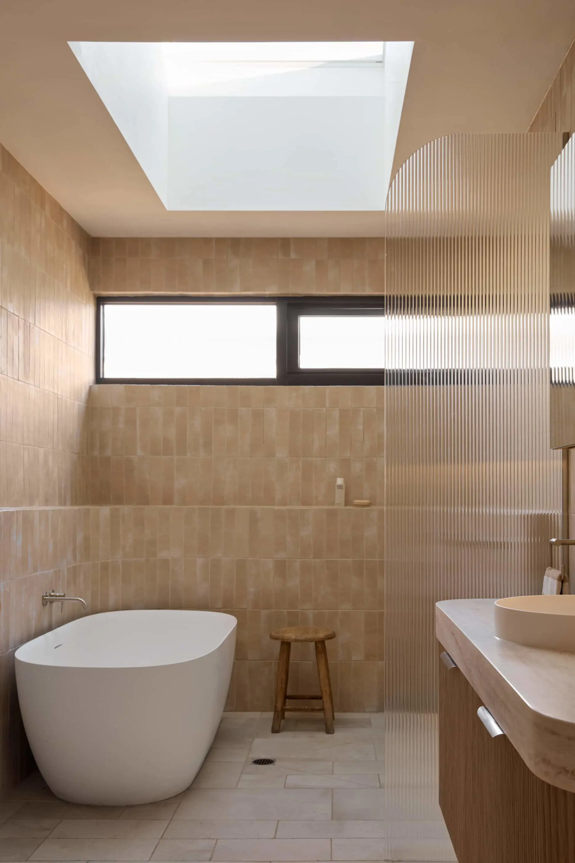Modern bathroom with beige tiled walls, white freestanding tub, wooden stool, skylight, and ribbed glass partition.