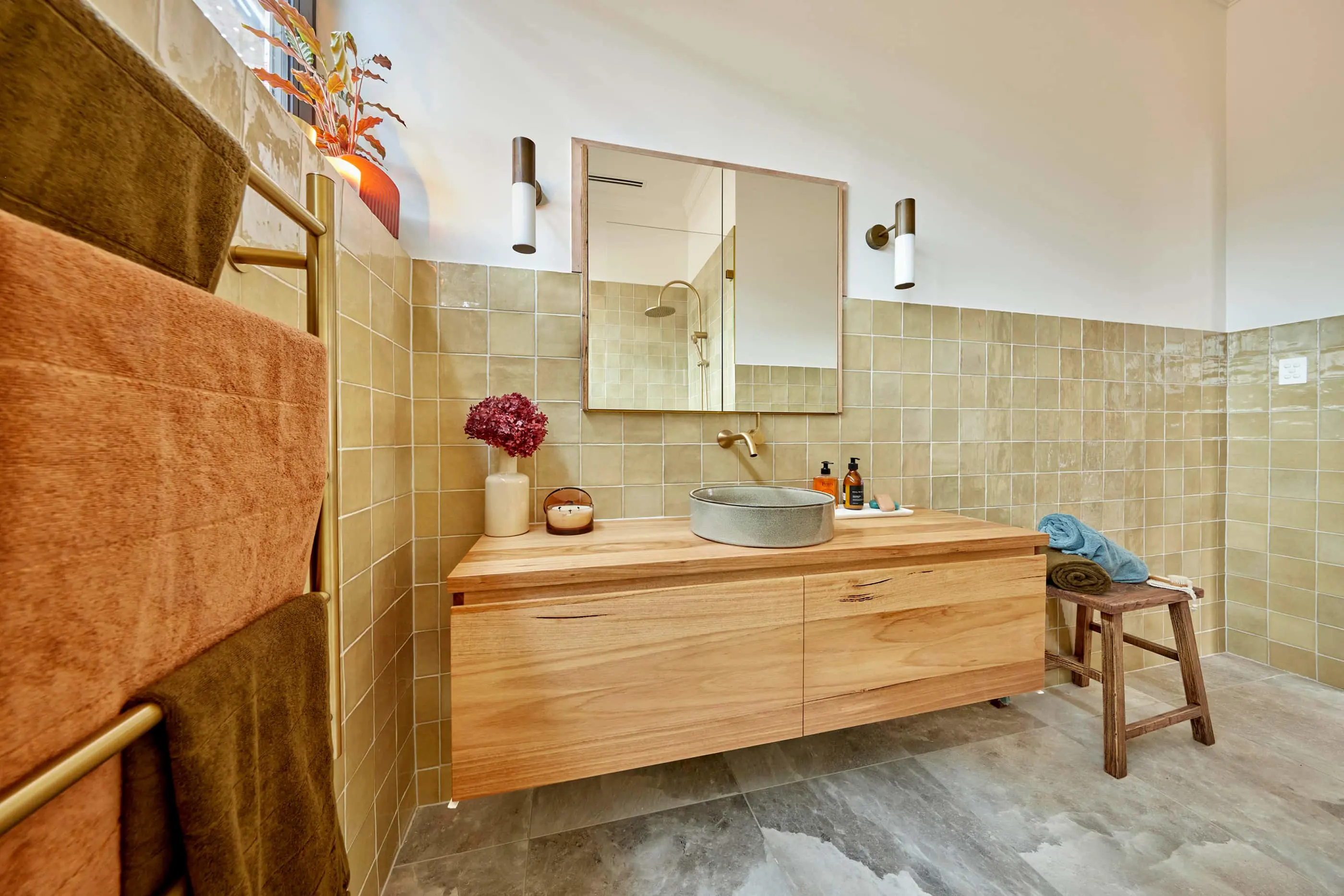 Modern bathroom with beige tiles, wooden vanity, vessel sink, mirror, wall sconces, and folded towels on a small stool.
