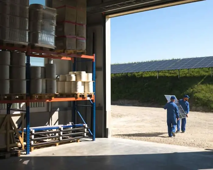 Warehouse interior with storage shelves, looking out to workers carrying solar panels with solar farm visible outside