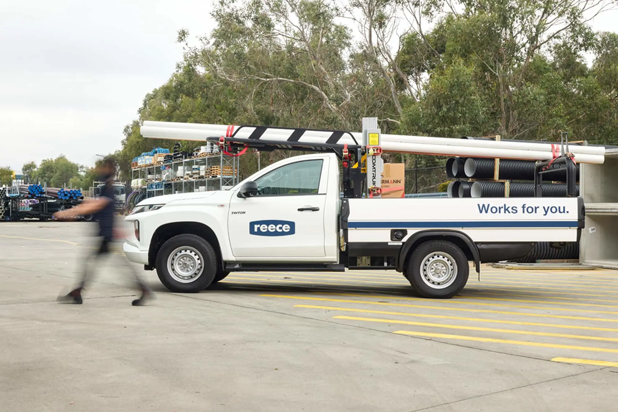 White Reece utility truck loaded with pipes and construction materials parked in a work yard with trees in background.