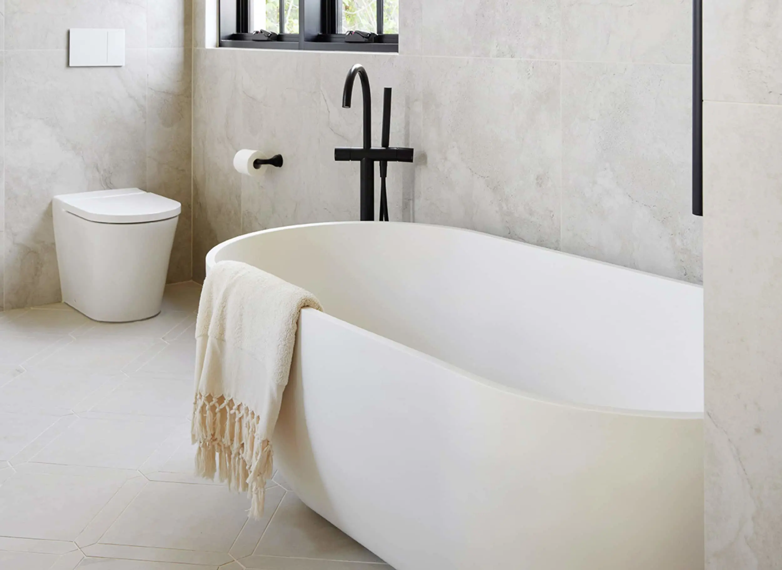 Modern bathroom with white freestanding tub, black fixtures, cream towel with tassels, and marble-look walls.