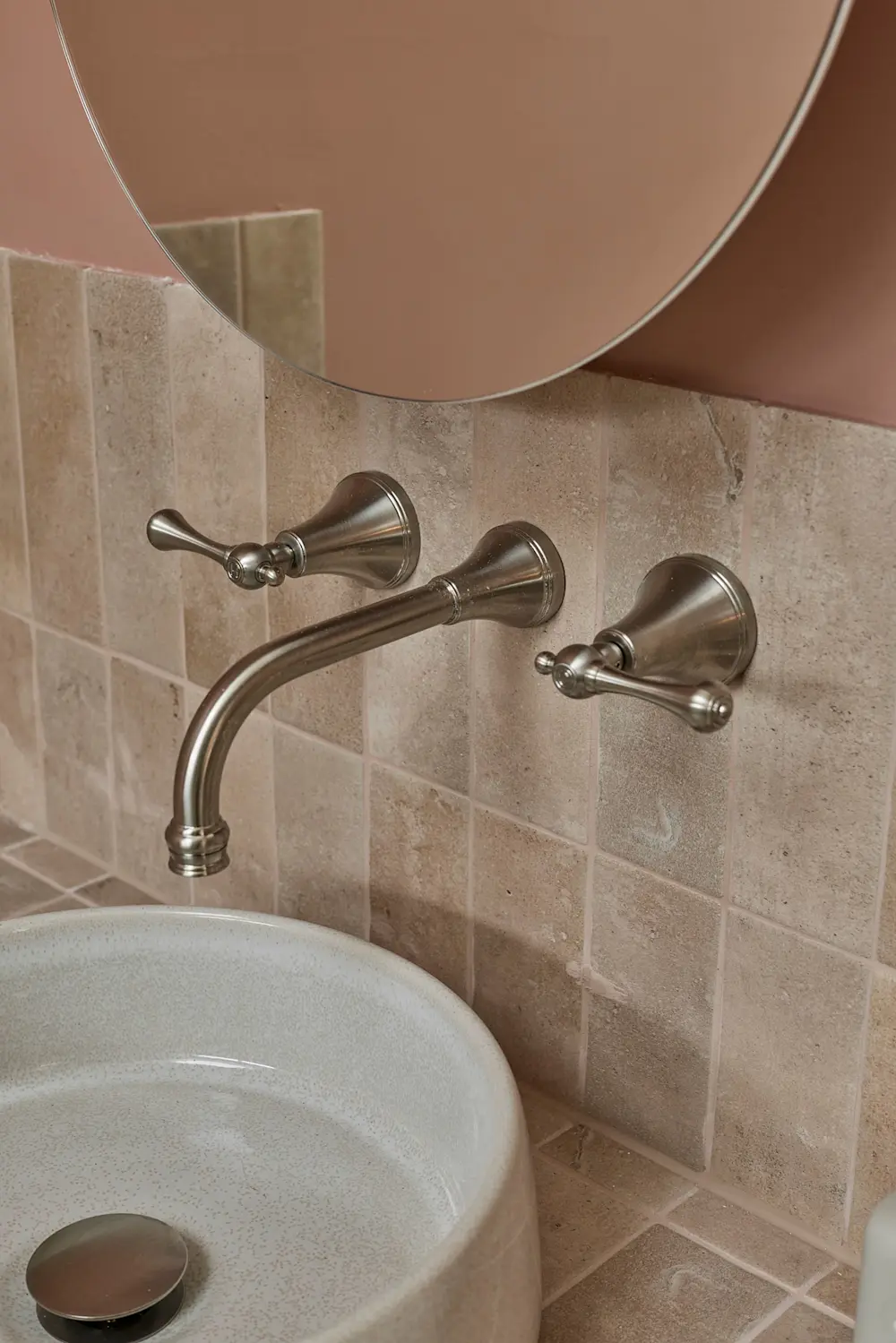 Wall-mounted brushed nickel bathroom faucet with dual handles above a white sink, beige tiled wall, and round mirror.