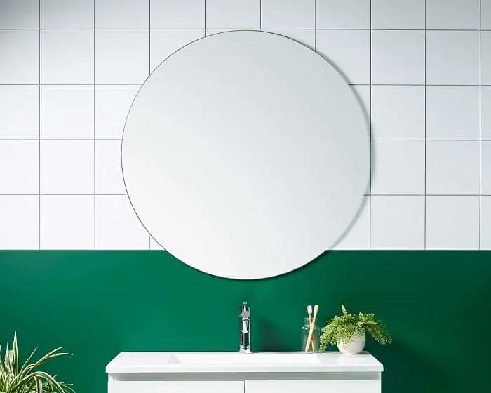Round mirror above bathroom sink with white tiles and green wall, featuring small plants and toothbrushes.
