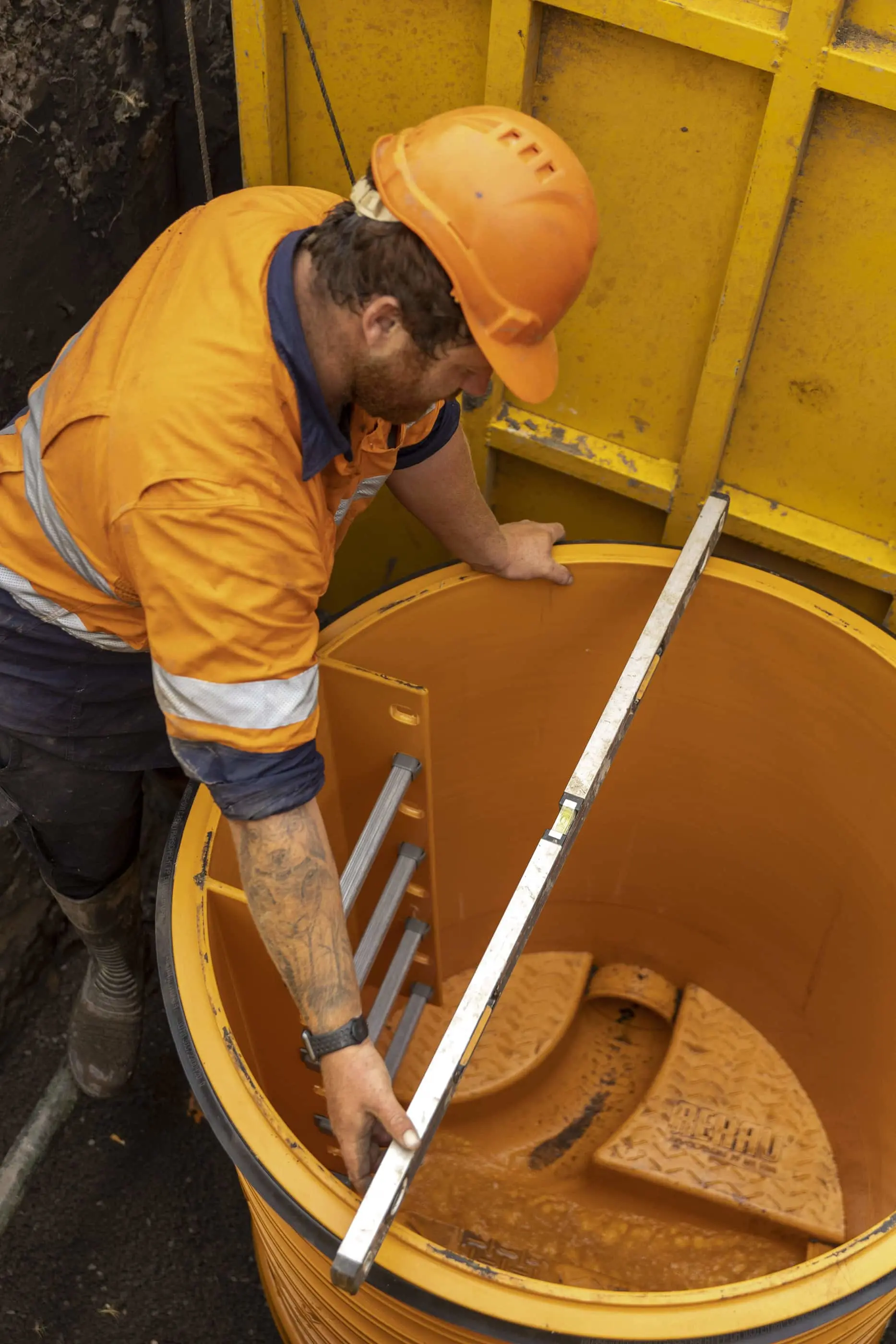 Construction worker in orange safety gear inspecting a yellow manhole or access chamber with ladder rungs.