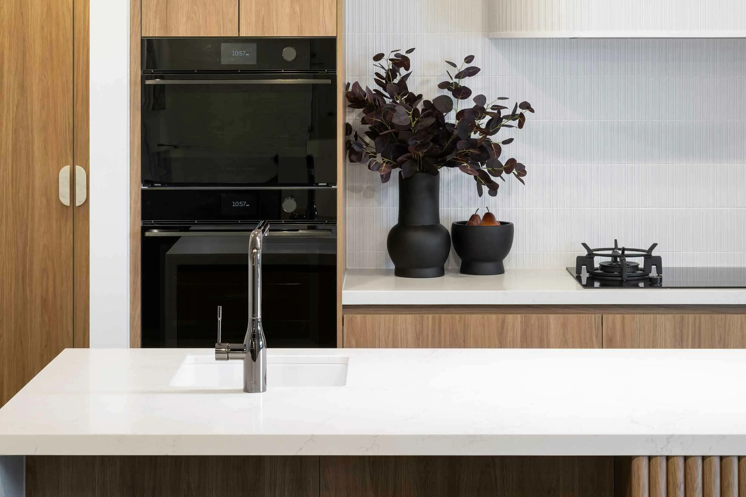 Modern kitchen with white countertops, built-in black ovens, wooden cabinets, and black decorative vases with plants.