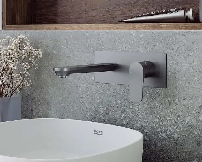 Modern wall-mounted gray faucet with flat spout above a white Roca sink, against textured gray stone wall with dried flowers.