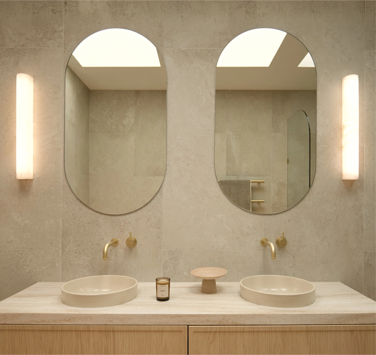 Modern bathroom with two oval mirrors above white vessel sinks on a marble countertop with brass faucets and wall sconces.