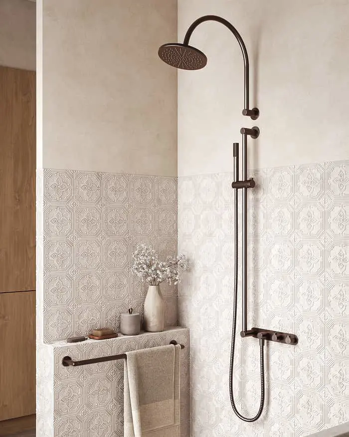 Bronze shower system with overhead rain head and handheld on patterned cream tiles, with a towel bar and small shelf nearby.