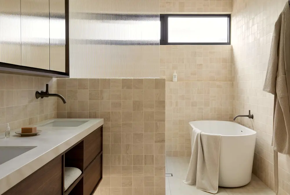Modern bathroom with beige tile walls, white freestanding tub, wooden vanity, and high window with textured glass.
