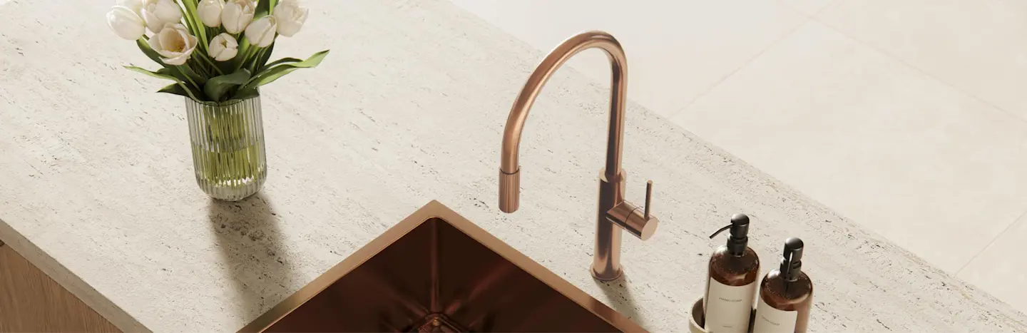Brushed bronze kitchen sink and faucet on a stone countertop, with white tulips in a vase and soap dispensers nearby.