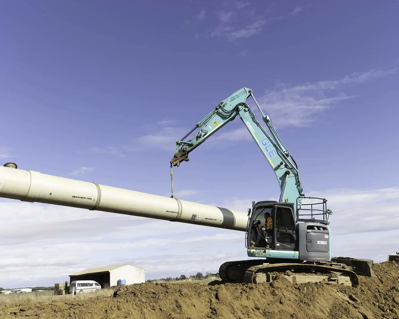 Blue excavator lifting a large white pipeline section at a construction site under clear blue sky.