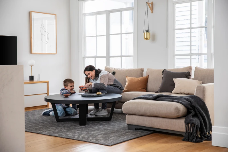 Woman and child playing together in bright living room with beige sectional sofa, black coffee table, and large windows.