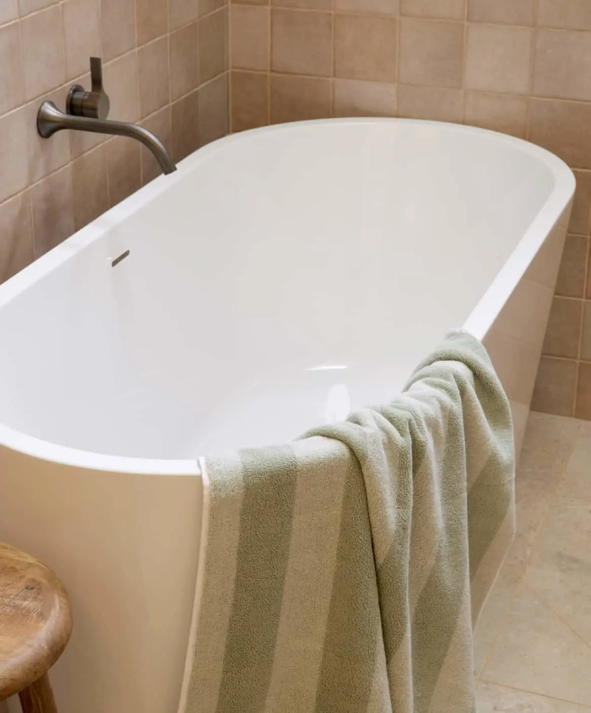 Modern white freestanding bathtub with wall-mounted faucet, light green towel draped over edge, and beige tiled walls.