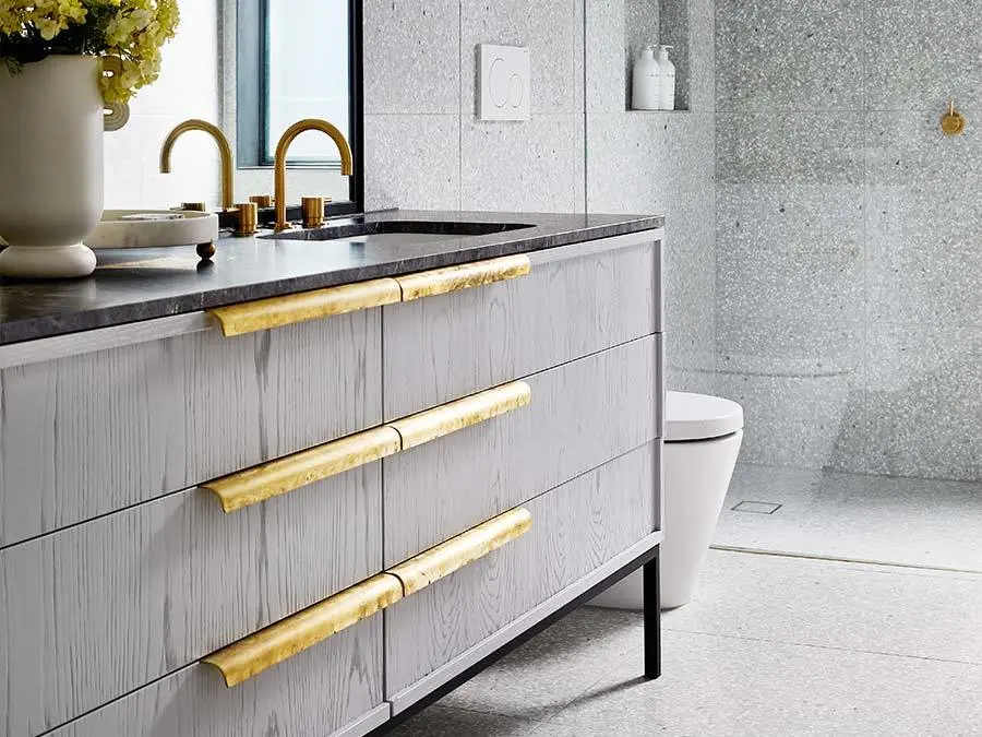 Modern bathroom with gray vanity featuring gold handles, brass faucet, dark countertop, and stone tile walls.