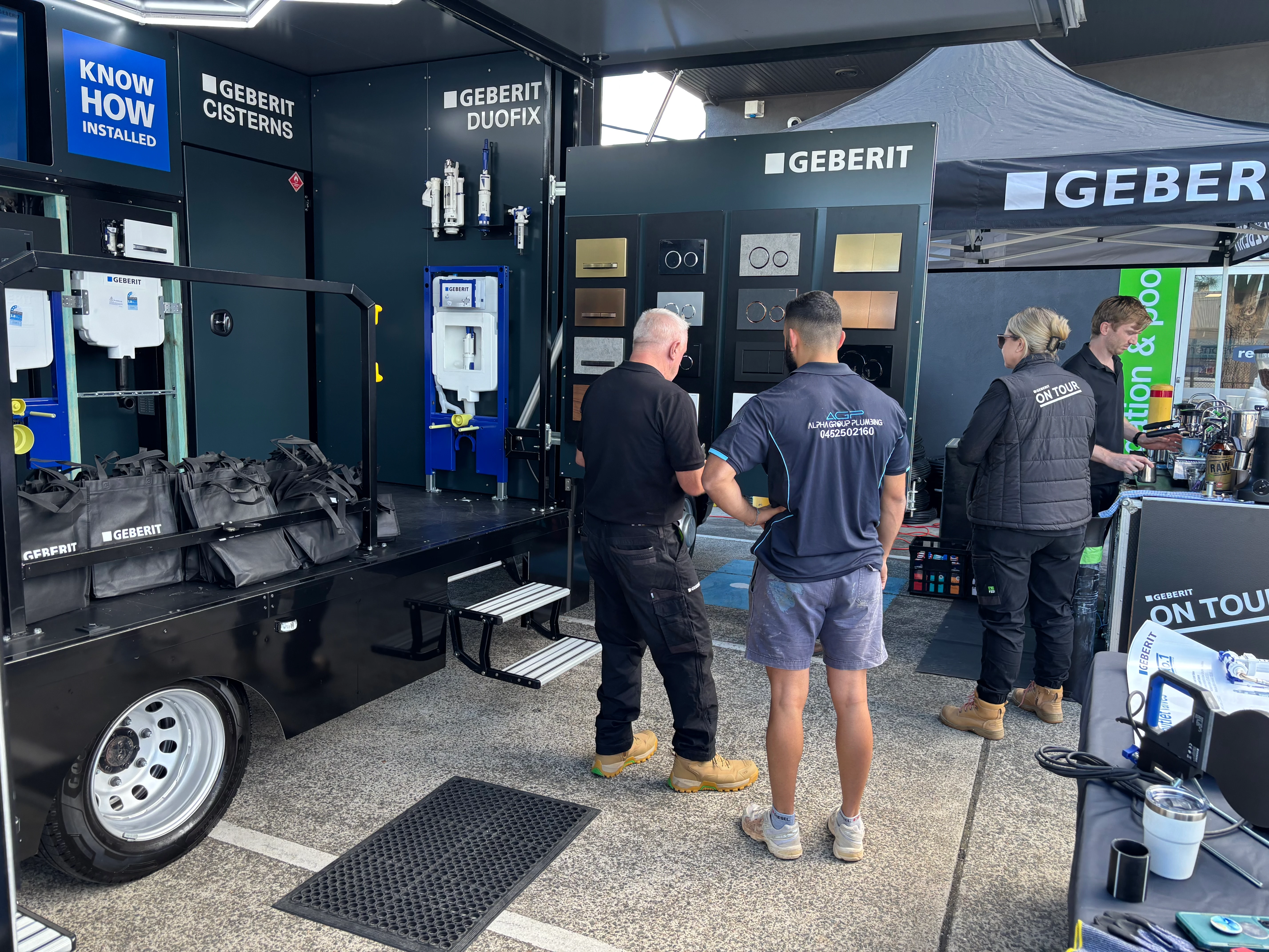 Four people stand at a Geberit outdoor trade display trailer showing cisterns, Duofix frames, and flush plate samples.