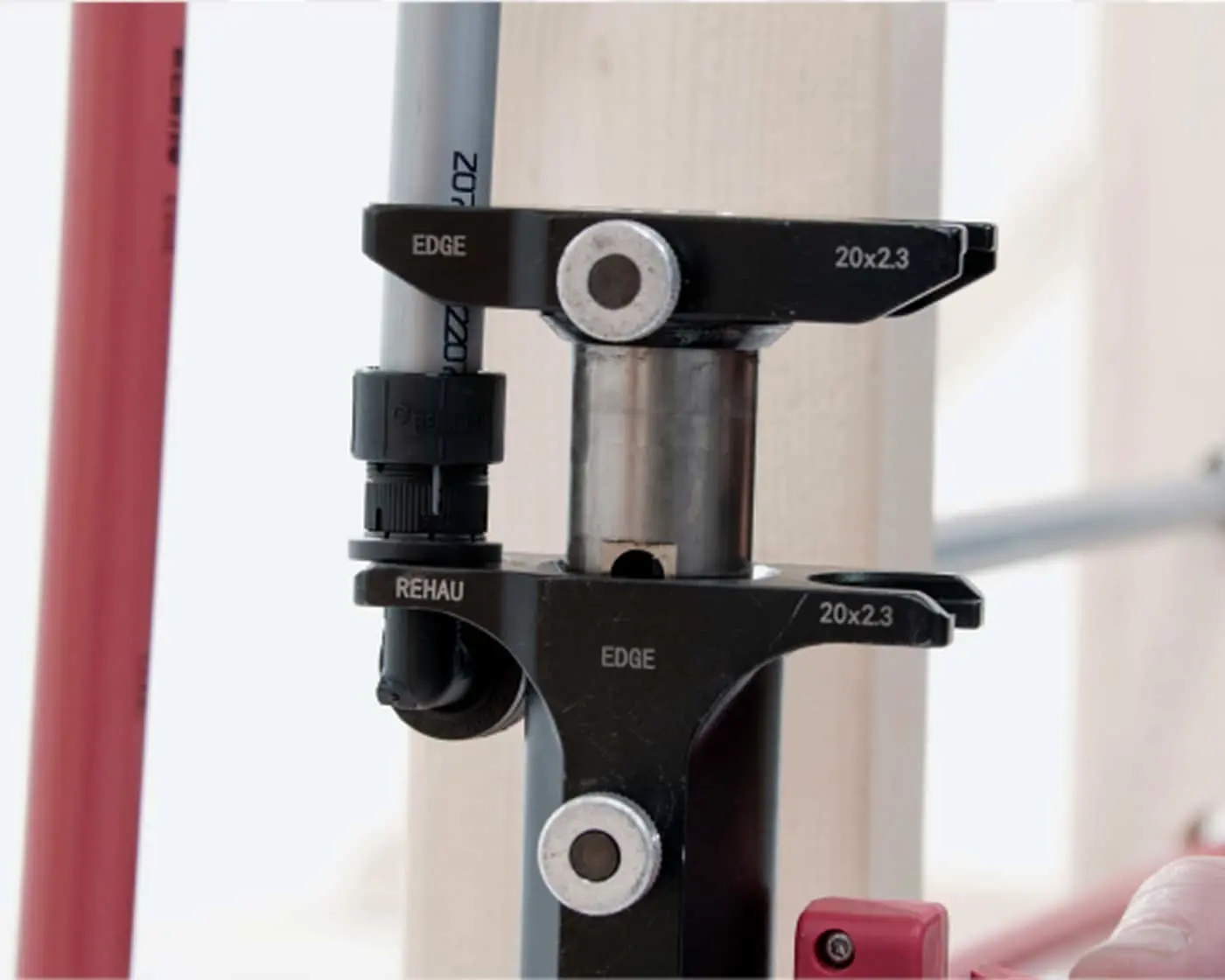 Black metal pipe fitting connector labeled "EDGE 20x2.3" with silver bolts attaching to gray and red pipes.