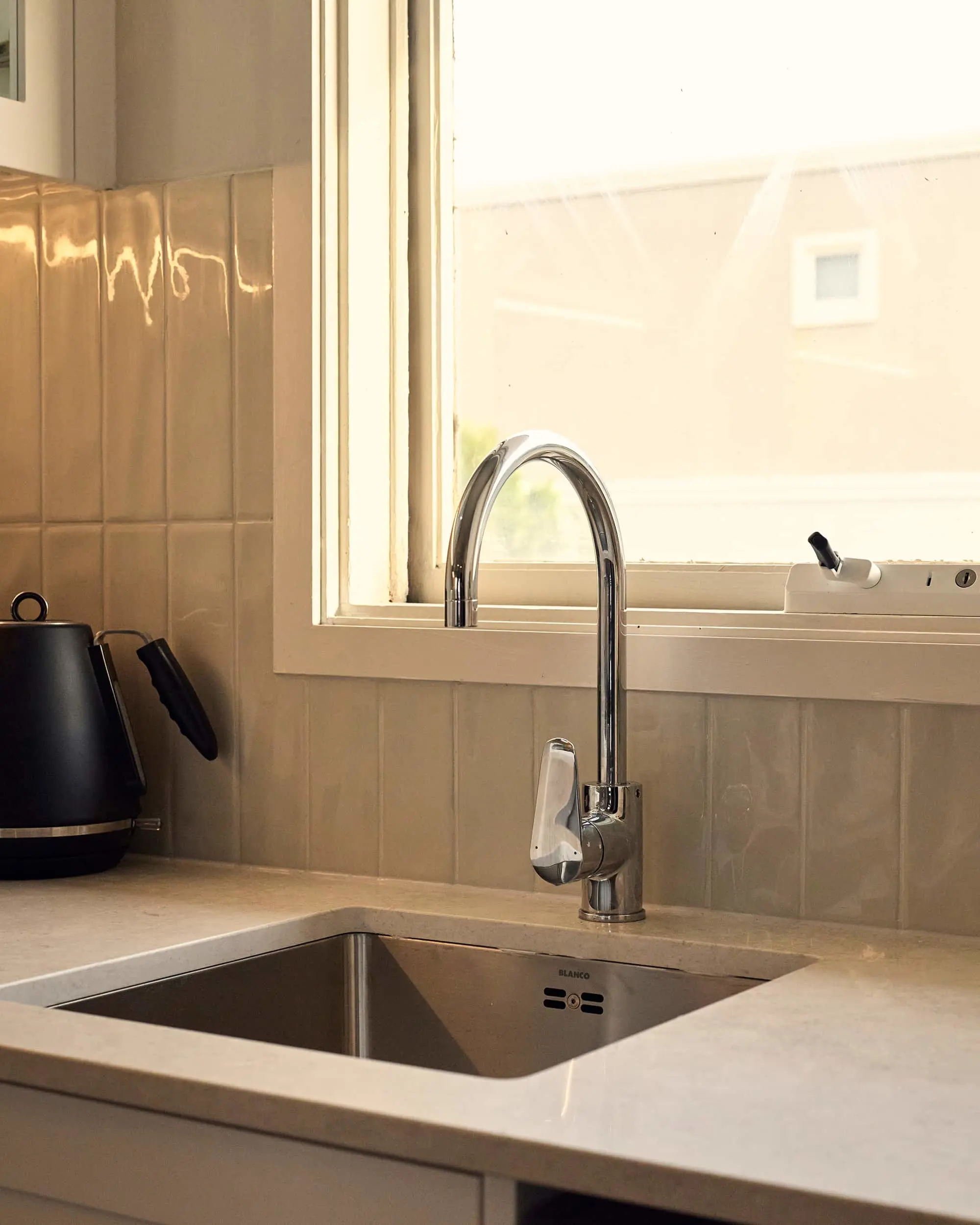 Chrome kitchen faucet over stainless steel sink with white countertop, window above, and black kettle to the left.