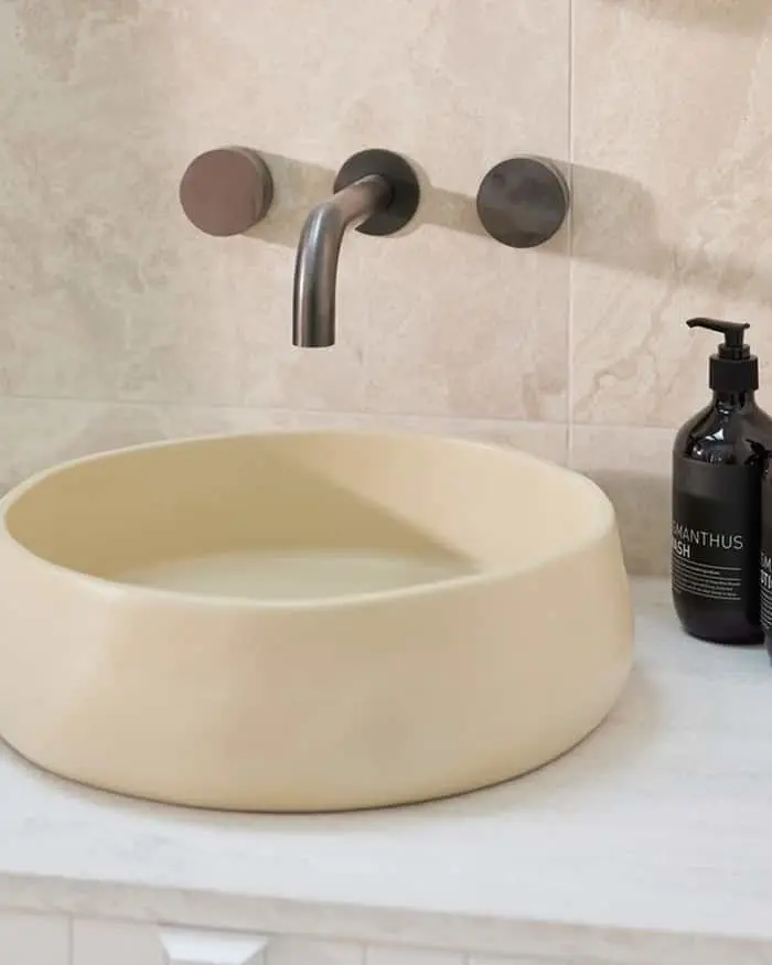 Modern bathroom sink with cream vessel basin, wall-mounted bronze faucet, and black soap dispenser on marble counter.