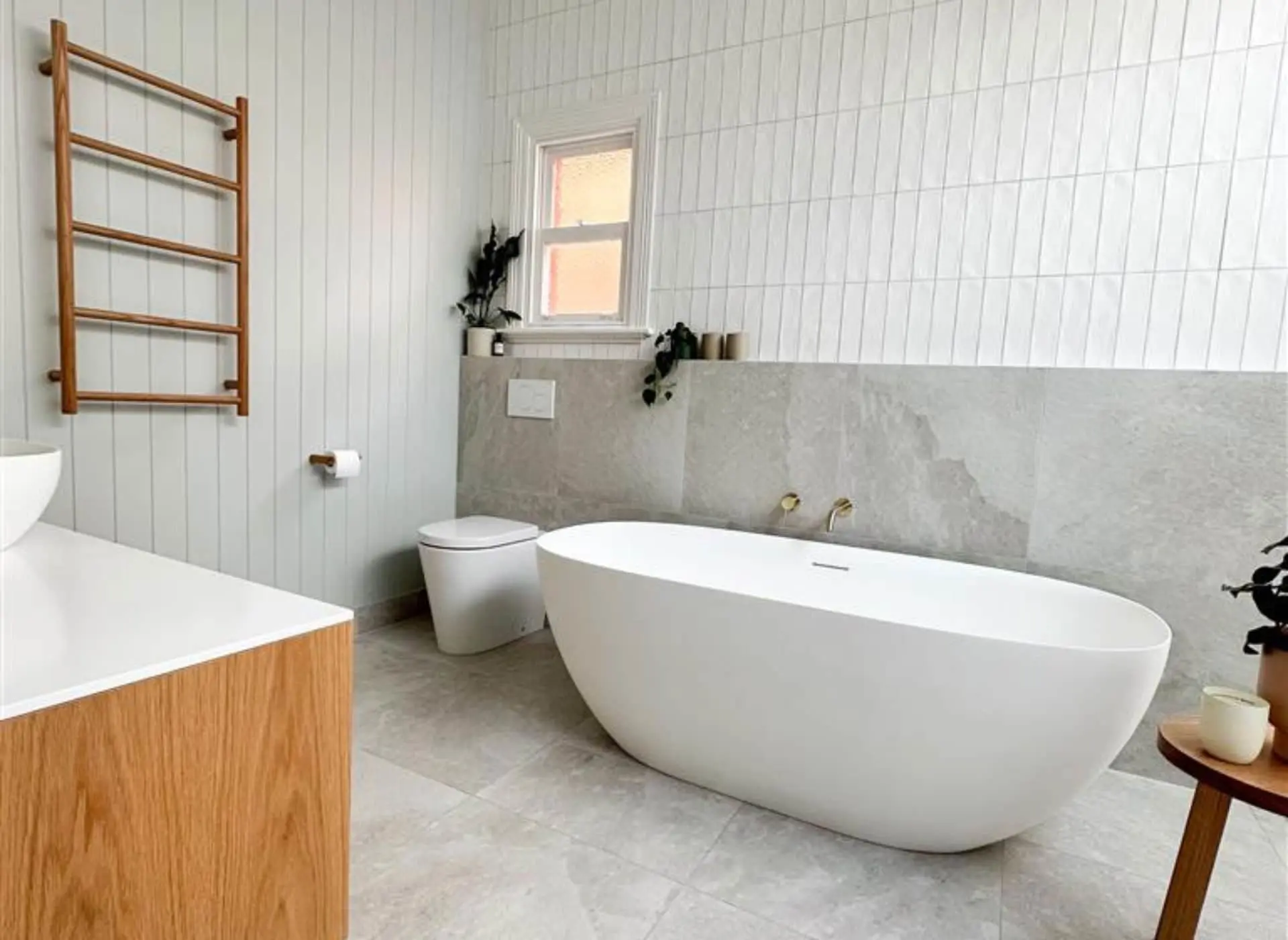 Modern bathroom with white freestanding tub, wooden towel rack, white toilet, and small window with potted plants.