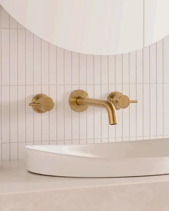 Modern bathroom with brass wall-mounted faucet and controls against white vertical tile, partial white sink visible.