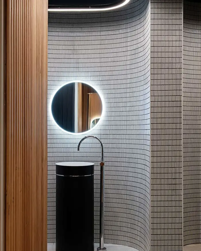 Modern bathroom with curved tile walls, illuminated round mirror, black pedestal sink, and wooden accent wall.