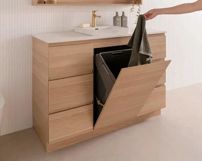 Modern wooden bathroom vanity with white sink and built-in laundry hamper, hand placing towel inside open drawer.