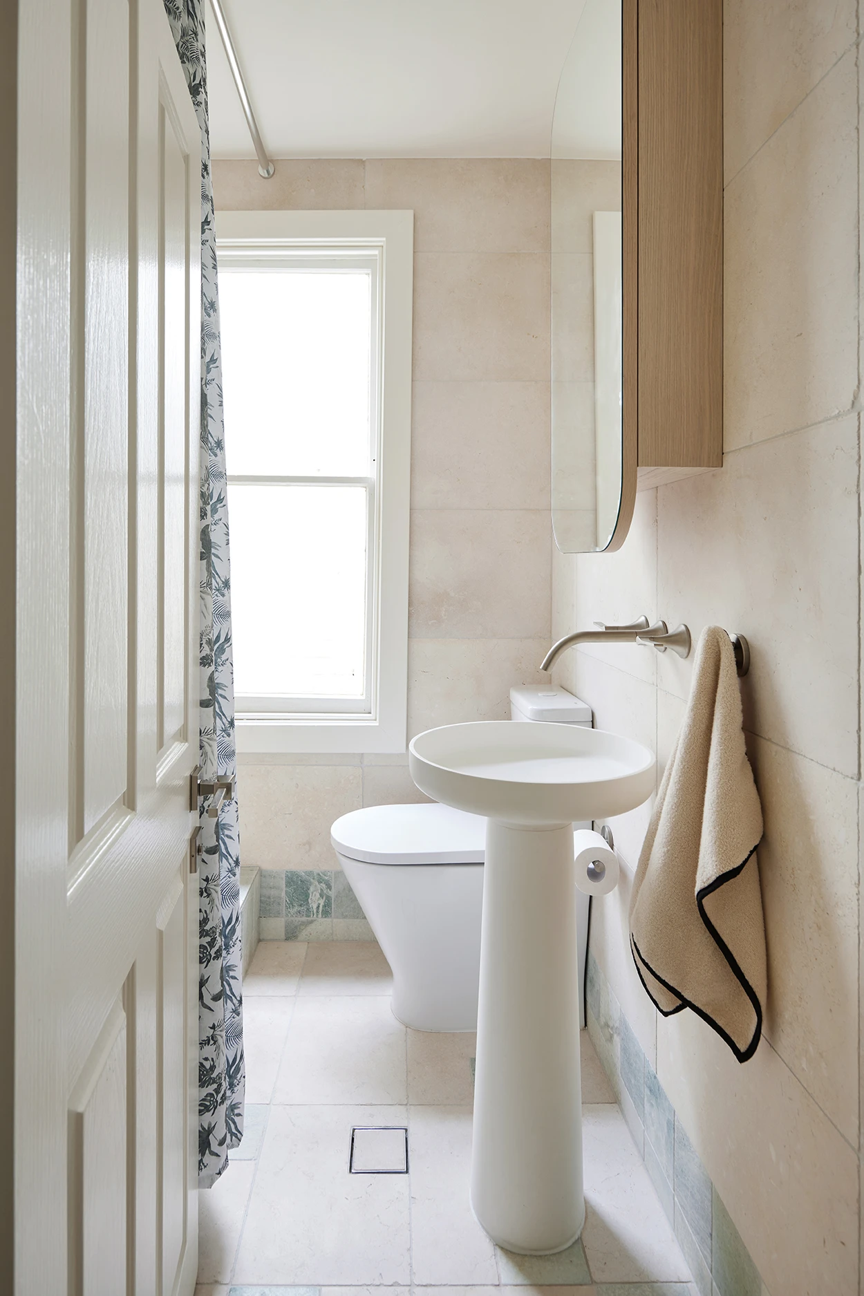 Narrow bathroom with pedestal sink, wall-mount faucet, toilet, floral shower curtain, beige stone tiles, and a wood cabinet above a mirror.