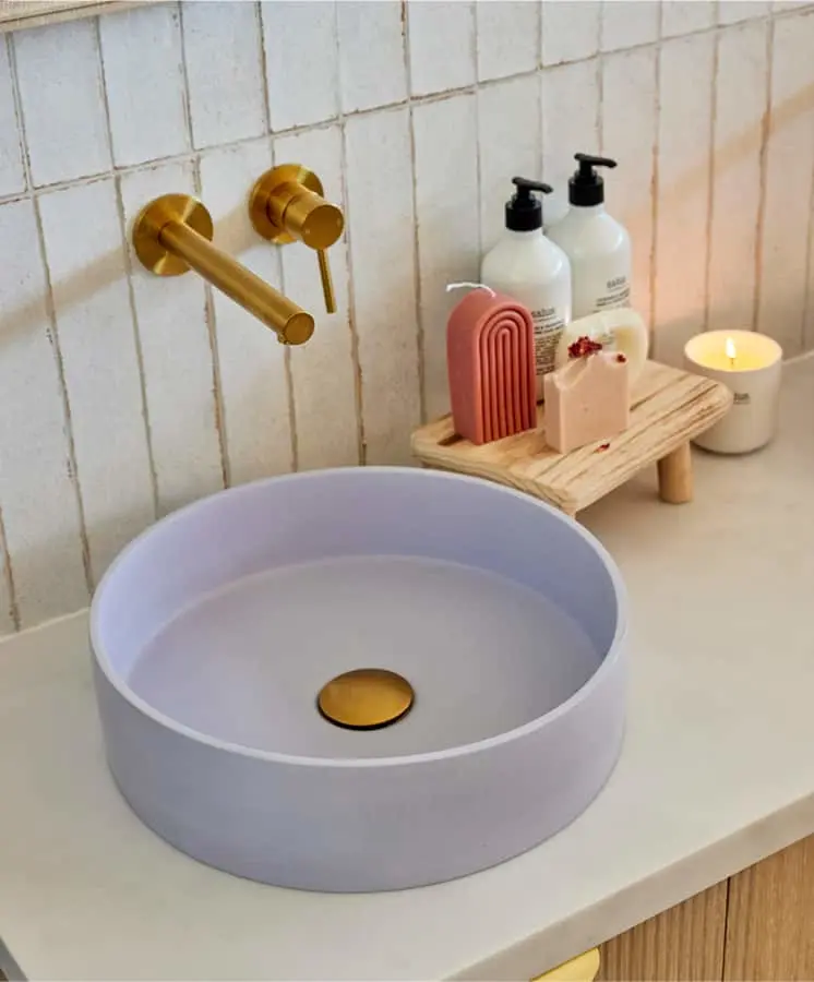 Lavender circular sink with gold fixtures on white countertop, wooden shelf with soap dispensers and candle nearby.