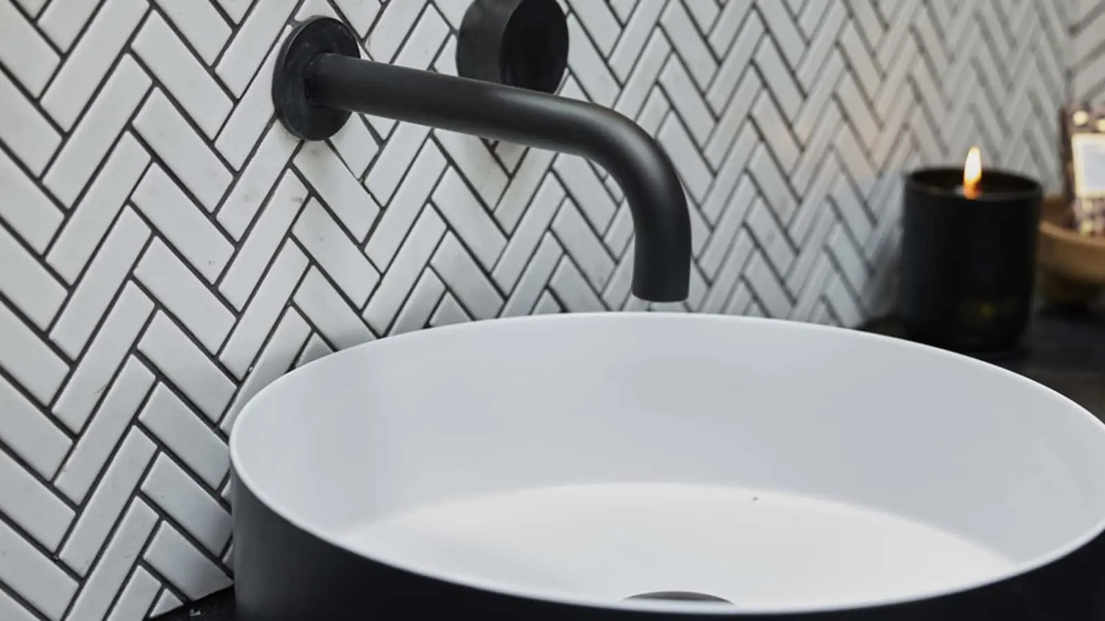Modern bathroom sink with black faucet against white herringbone tile wall, lit candle in background.