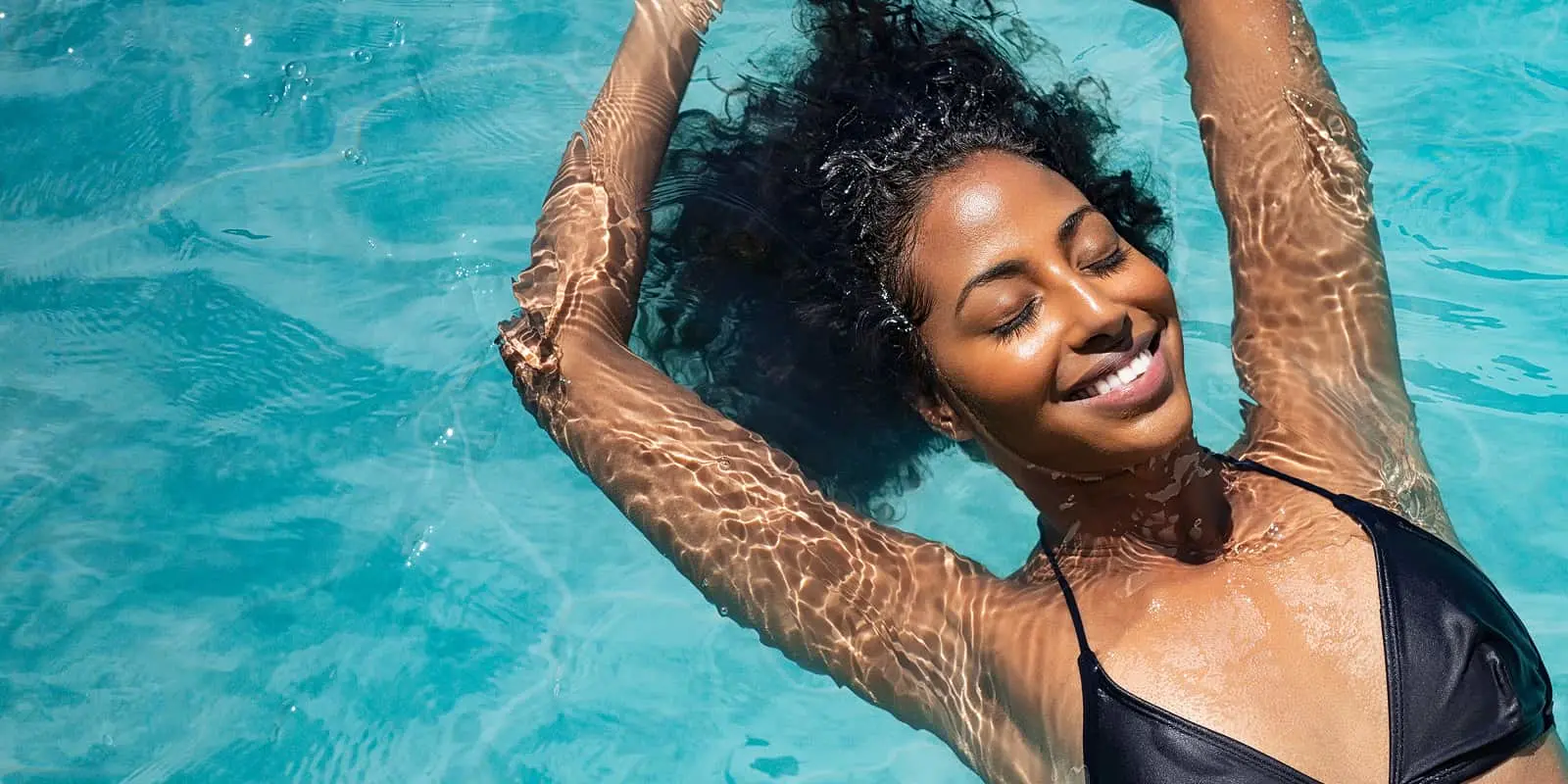 Person with curly hair floating in clear turquoise water, wearing a black swimsuit, eyes closed with a peaceful smile.