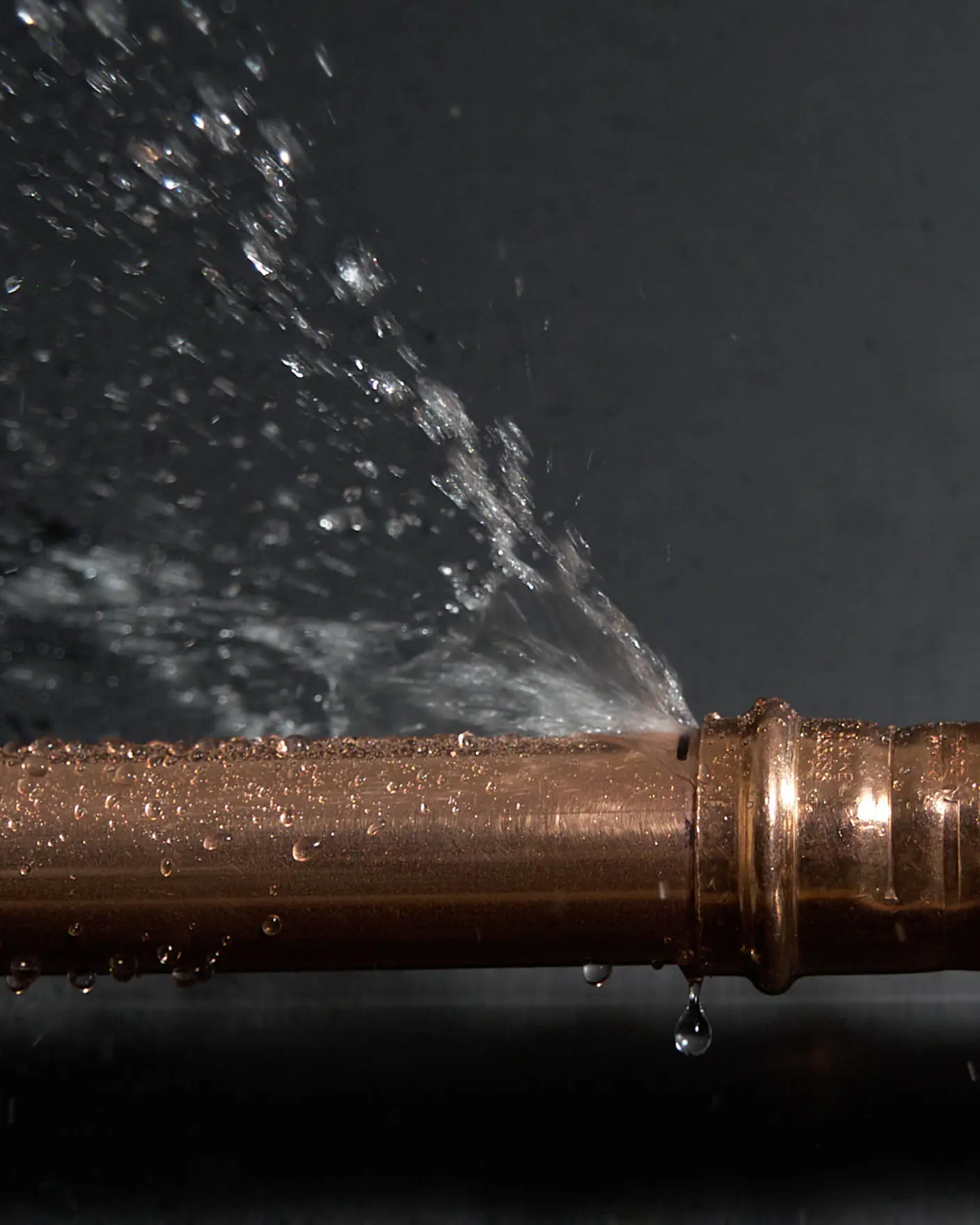 Water spraying from a leak in a copper pipe, with droplets visible against a dark background.