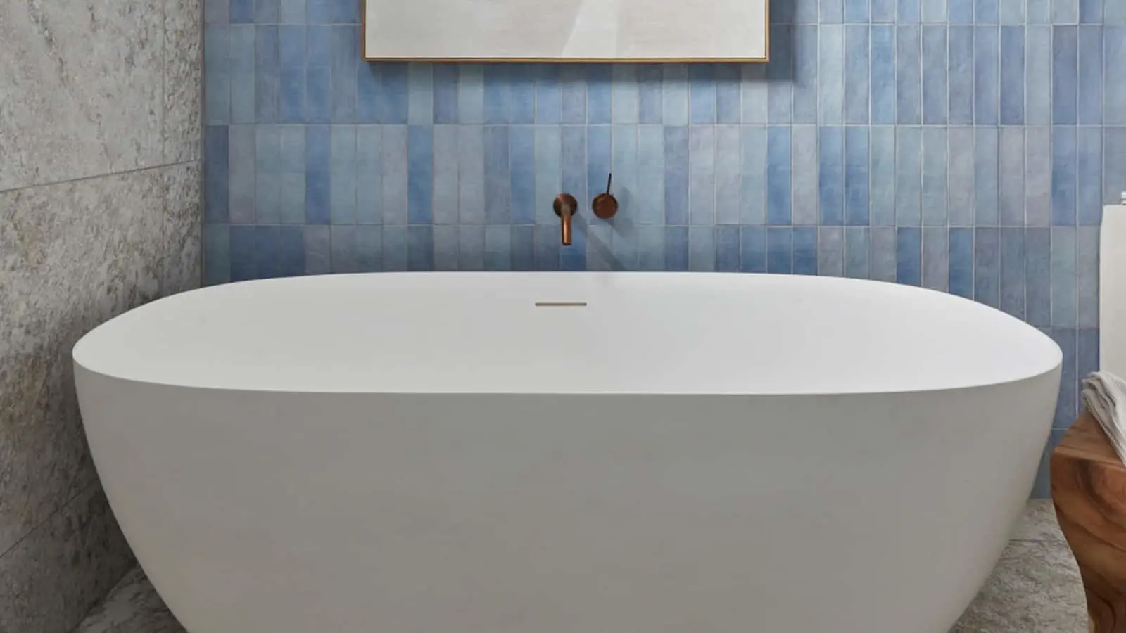Modern white freestanding bathtub against blue tiled wall with bronze wall-mounted faucet and framed artwork above.