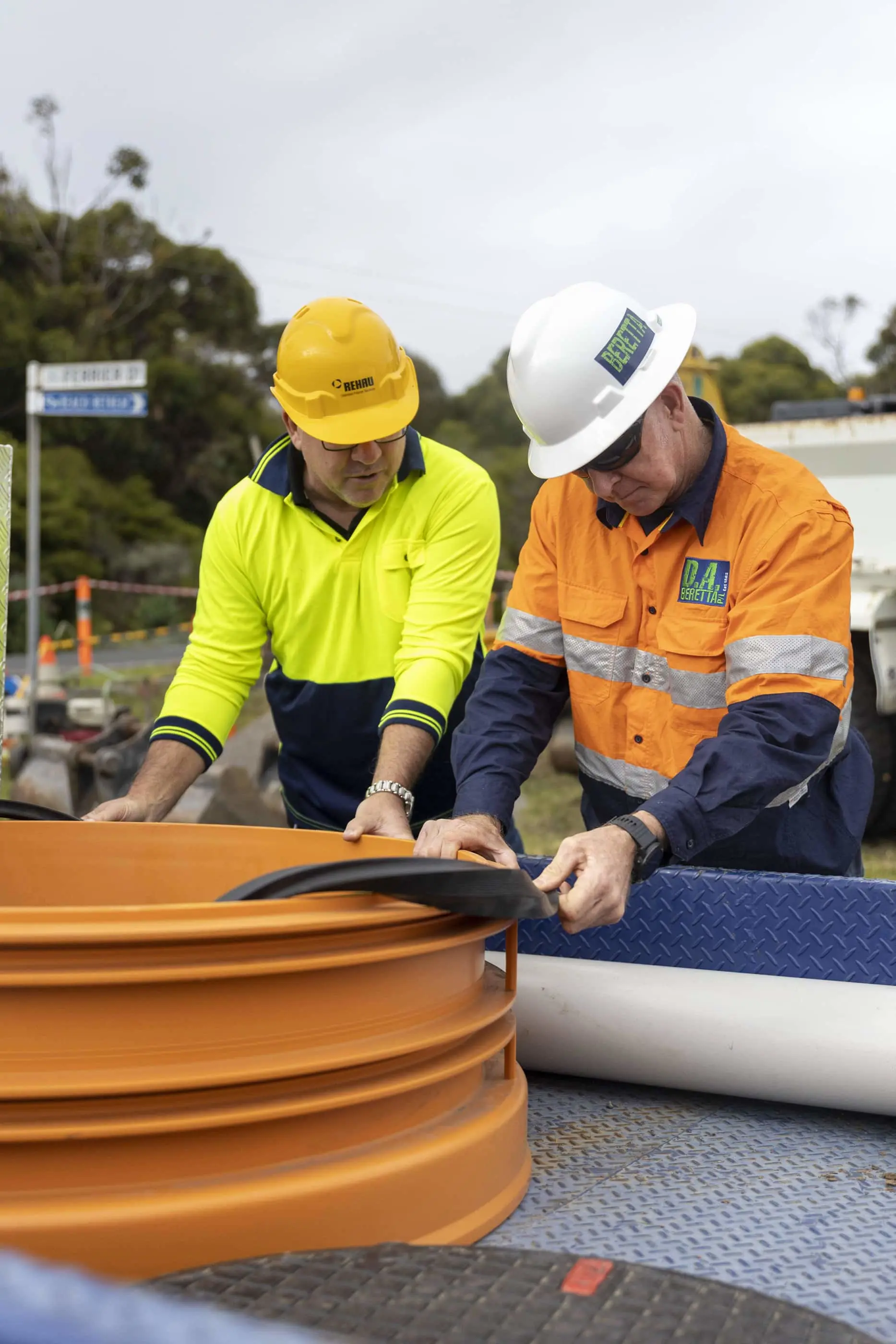Two construction workers in safety gear examining orange drainage pipes at an outdoor worksite.