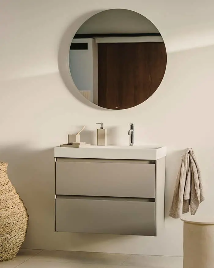 Modern bathroom with round mirror above wall-mounted gray vanity, chrome faucet, and woven basket nearby.