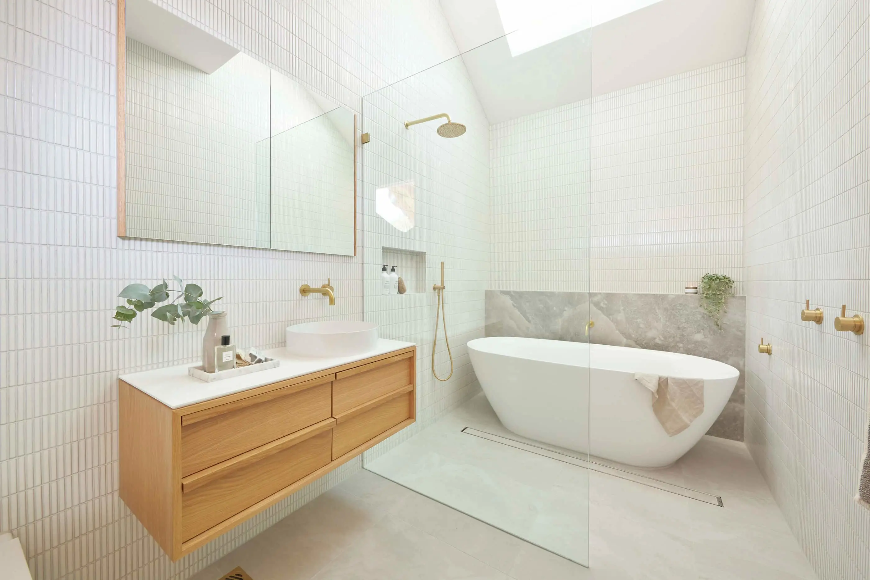 Modern white bathroom with freestanding tub, wooden vanity, gold fixtures, and skylight in minimalist design.