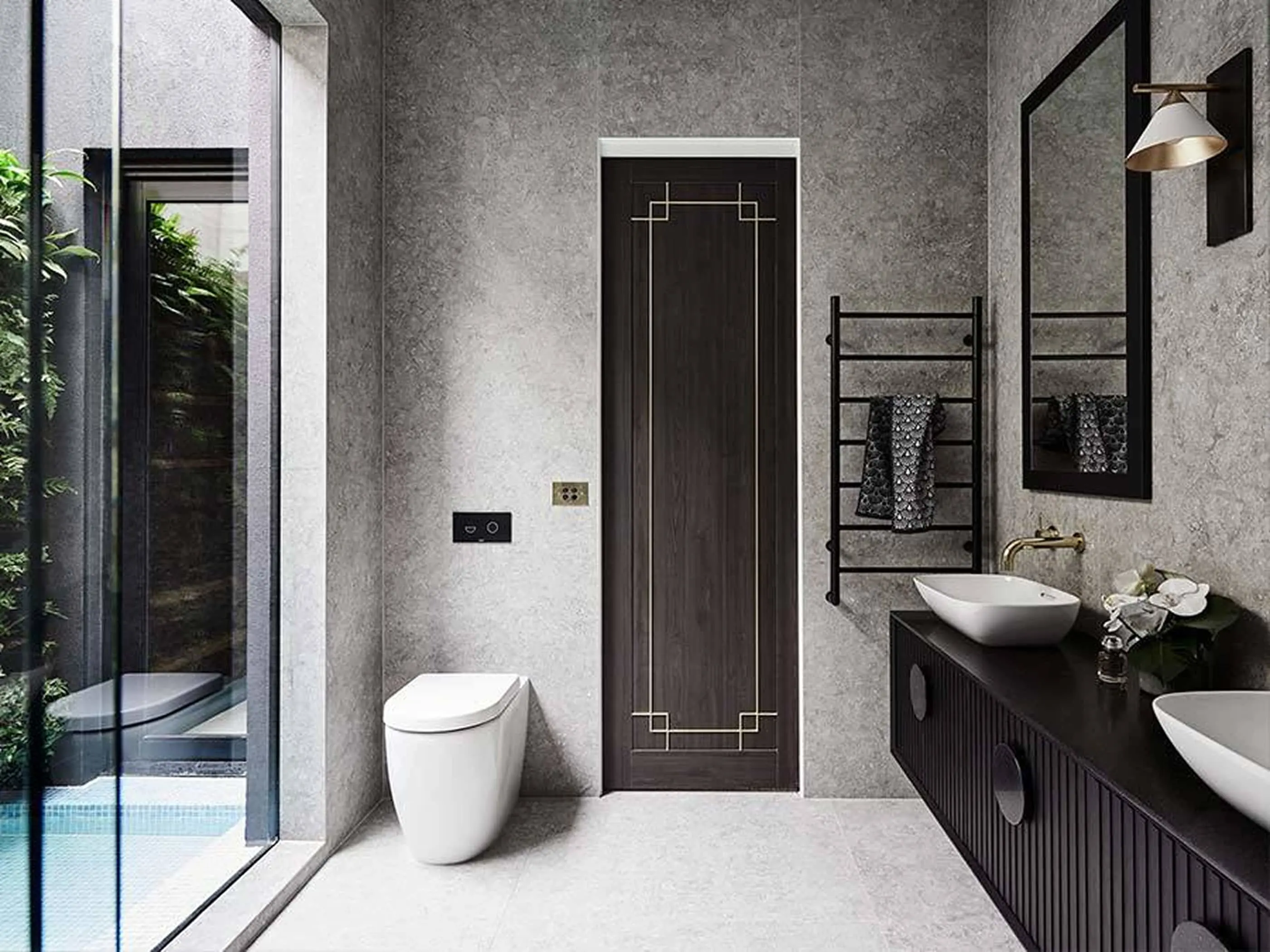 Modern bathroom with gray walls, dark wood door, white toilet, double sink vanity, and large window overlooking greenery.