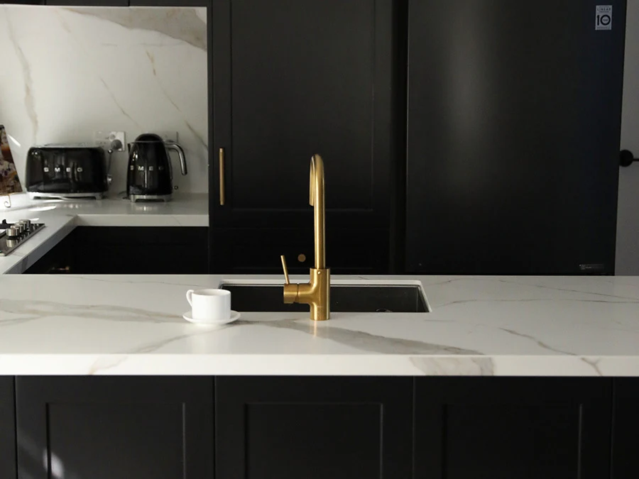 Modern kitchen with black cabinets, white marble countertops, gold faucet, and small coffee cup next to sink.